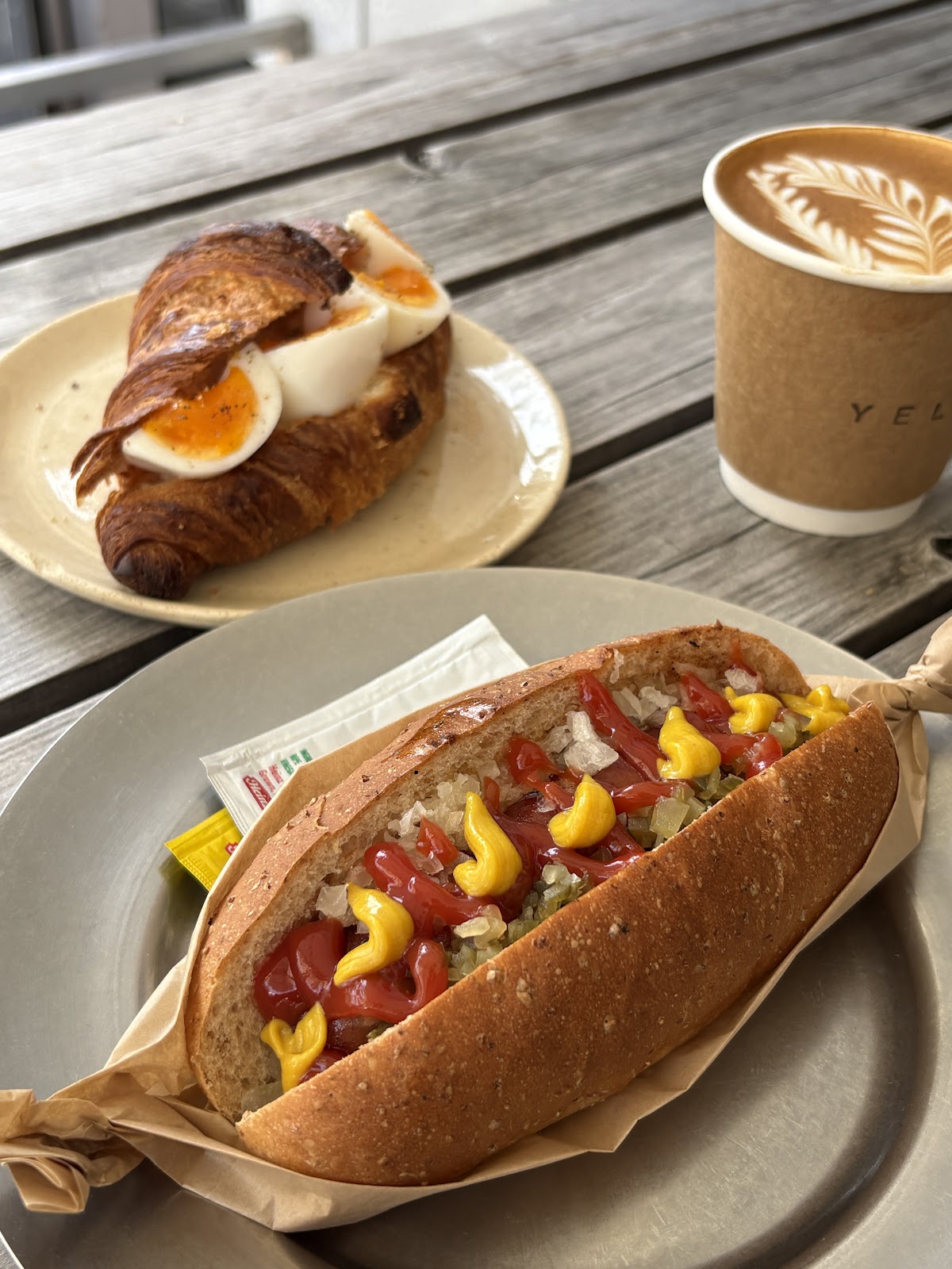 グレーの皿にケチャップとマスタードをかけたホットドッグ、奥に半熟卵のクロワッサンサンドとラテアートの紙カップ