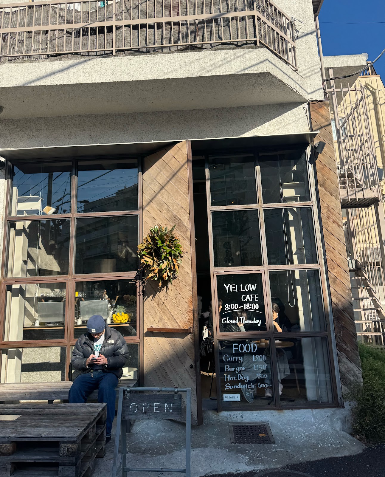 「YELLOW CAFE 8:00-18:00 Closed Thursday」と書かれたガラスの案内板と木製ドア、日差しの中で外のベンチに座る人